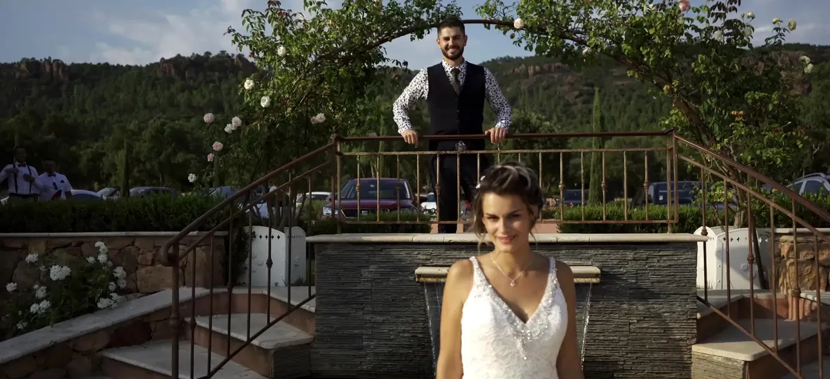 Christelle en robe de mariée devant une fontaine, Guillaume l'attend sous une arche de roses